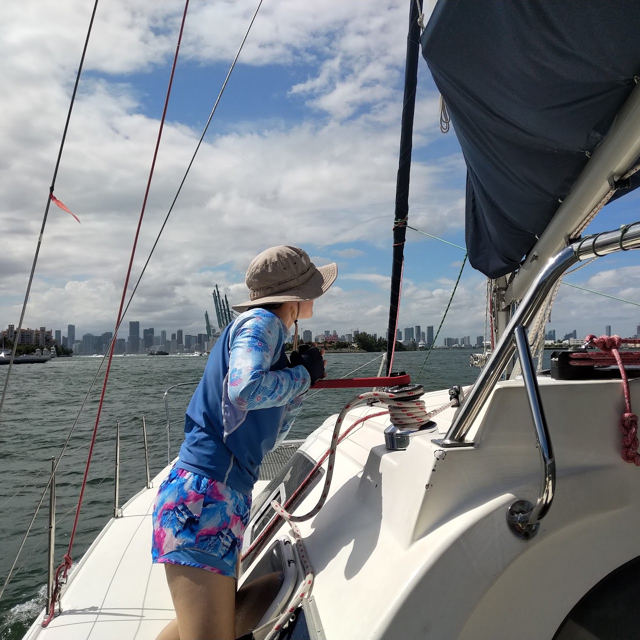 A woman wearing a floppy hat on a sailboat faces away as she turns a crank to tighten the mainsheet.