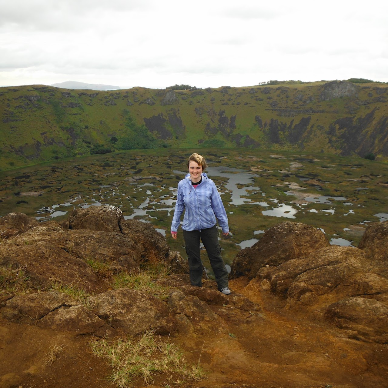 A woman in a light blue raincoat stands in front of a volcanic crater full of swampy vegetation on Rapa Nui.