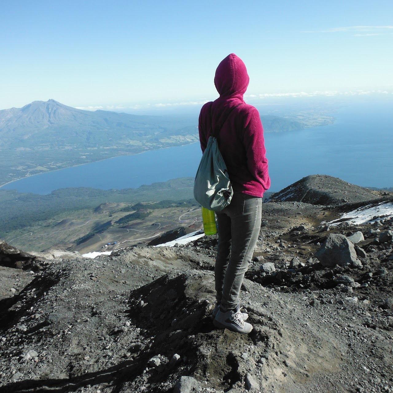 A woman in a hooded magenta sweatshirt standing on volcanic rock looks out over a lake from a high vantage point.