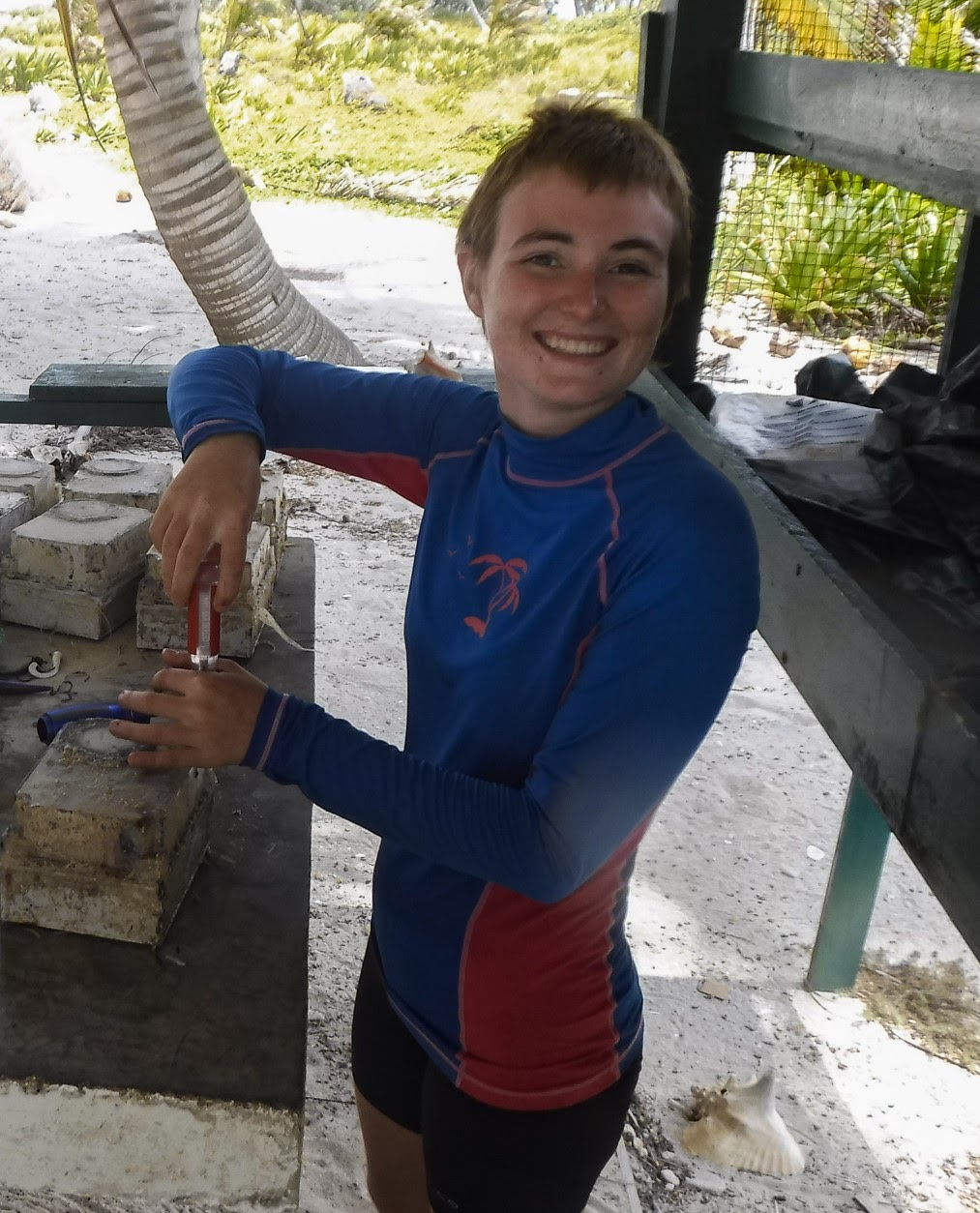 Stephanie, a young woman with short brown hair and a deep tan, smiles at the camera as she fixes an algae-covered camera housing in a wooden shelter.